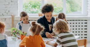 A teacher guides children around a table in a bright, engaging classroom setting.